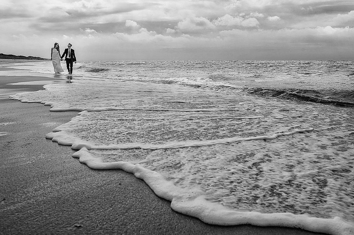 Ein Hochzeitspaar spaziert Hand in Hand und barfuss direkt auf dem Ufer am Strand in Kampen auf Sylt.