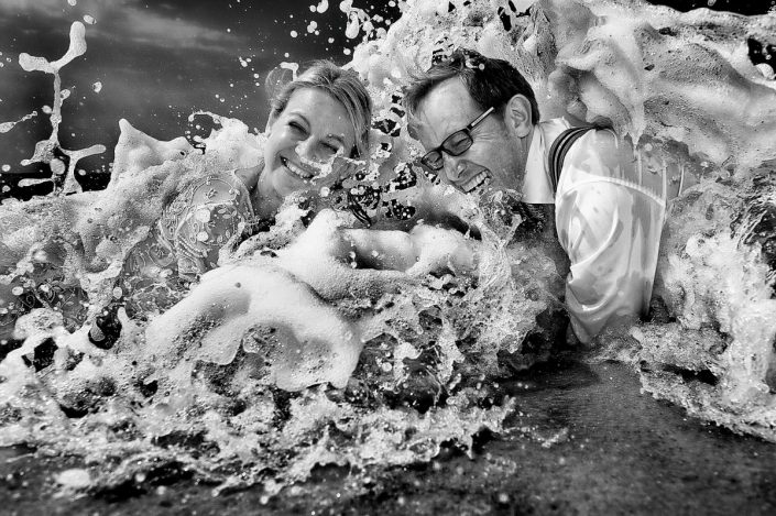 Vollgekleidetes Hochzeitspaar liegt im Wasser am sylter Strand und herzlich lacht, weil die gerade von der Welle getroffen sind und komplett nass sind.